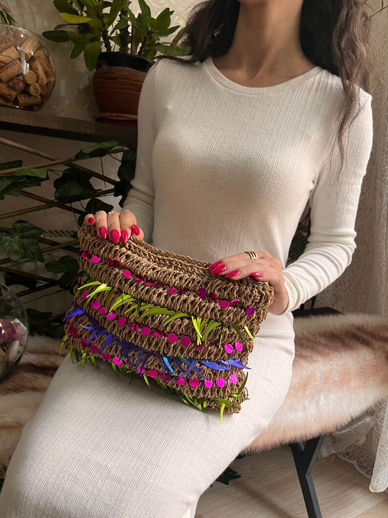 May include: A woven brown clutch with neon green, pink, and blue accents. The rectangular bag is held by a person wearing a white long-sleeved dress. The clutch features a textured design with colorful embellishments.