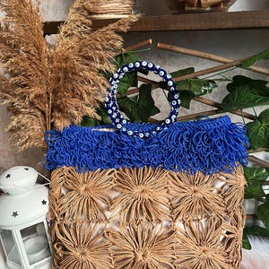 May include: A handcrafted tote bag with a natural woven base and a vibrant blue fringe top. The bag features a decorative handle adorned with blue and white beads. The bag's design incorporates a floral pattern, creating a unique and stylish accessory.