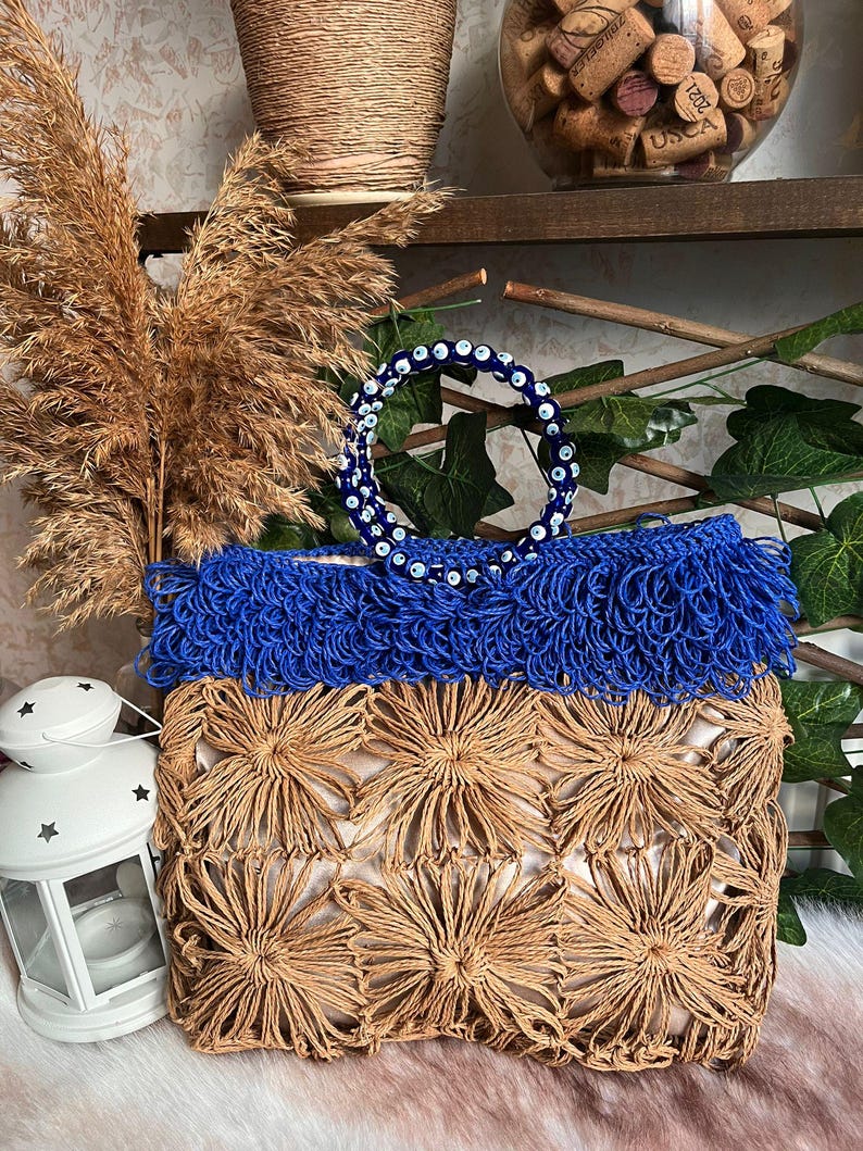 May include: A handcrafted tote bag with a natural woven base and a vibrant blue fringe top. The bag features a decorative handle adorned with blue and white beads. The bag's design incorporates a floral pattern, creating a unique and stylish accessory.