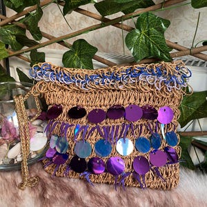 May include: A woven raffia clutch with a gold chain strap. The bag features a blue and white trim at the top and is embellished with iridescent purple sequins. The bag is rectangular and has a textured appearance.