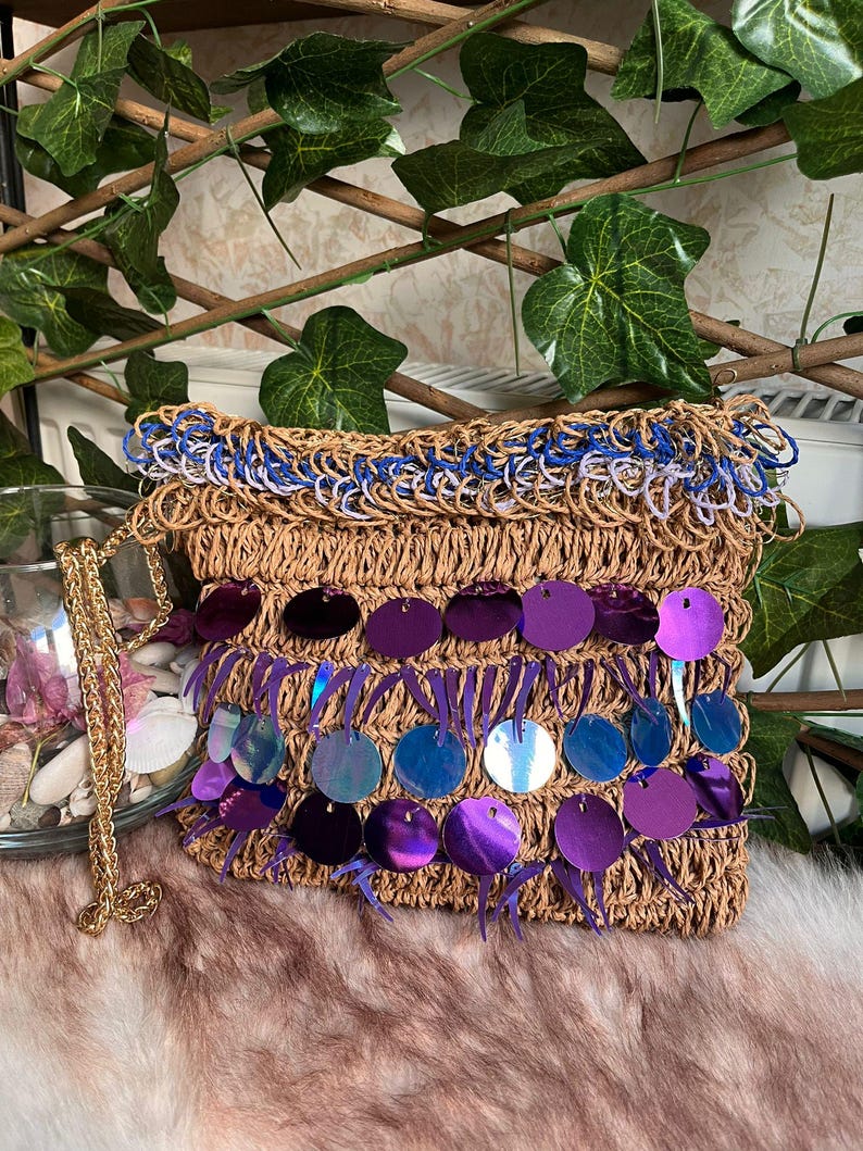 May include: A woven raffia clutch with a gold chain strap. The bag features a blue and white trim at the top and is embellished with iridescent purple sequins. The bag is rectangular and has a textured appearance.