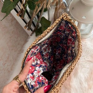 May include: A woven straw clutch bag with a gold zipper, revealing a floral patterned interior. The bag's exterior is a natural tan color, and the interior fabric features red and white flowers on a navy blue background.