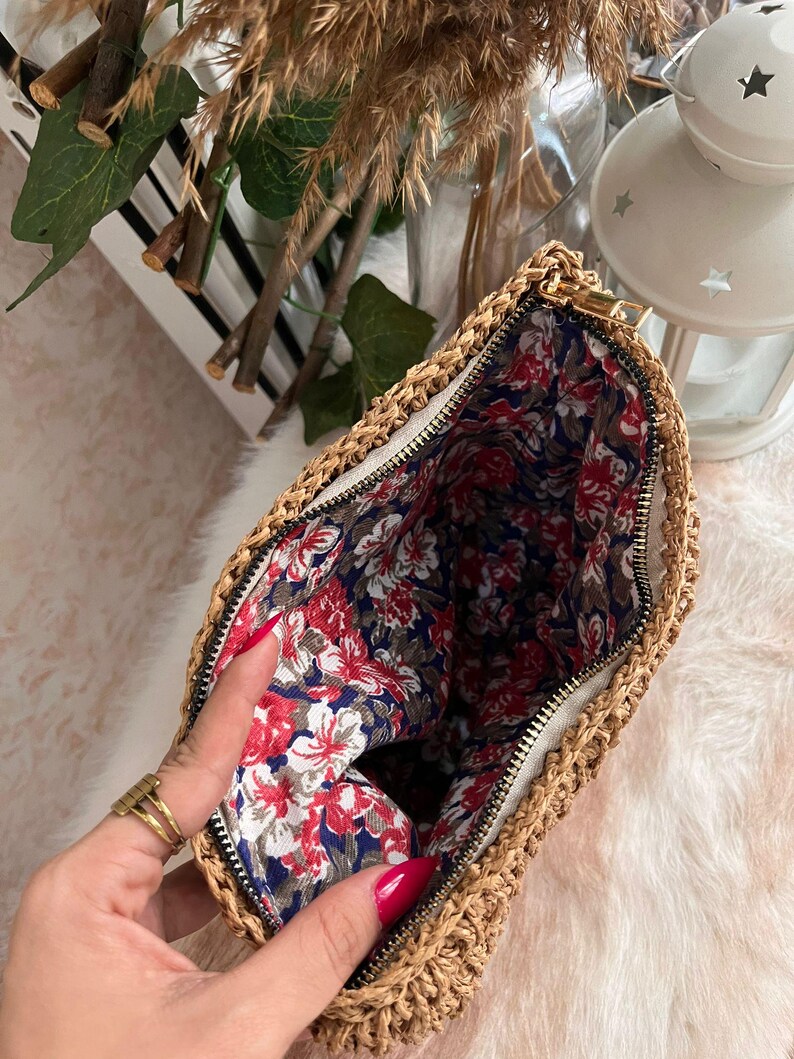 May include: A woven straw clutch bag with a gold zipper, revealing a floral patterned interior. The bag's exterior is a natural tan color, and the interior fabric features red and white flowers on a navy blue background.