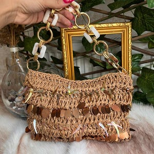 K&ouml;nnte beinhalten: Eine gewebte Raffia-Handtasche mit einem dekorativen Fransenbesatz aus Pailletten. Die Tasche hat einen goldfarbenen Kettenhenkel mit wei&szlig;en und braun marmorierten Akzenten. Die Tasche wird von einer Hand gehalten.