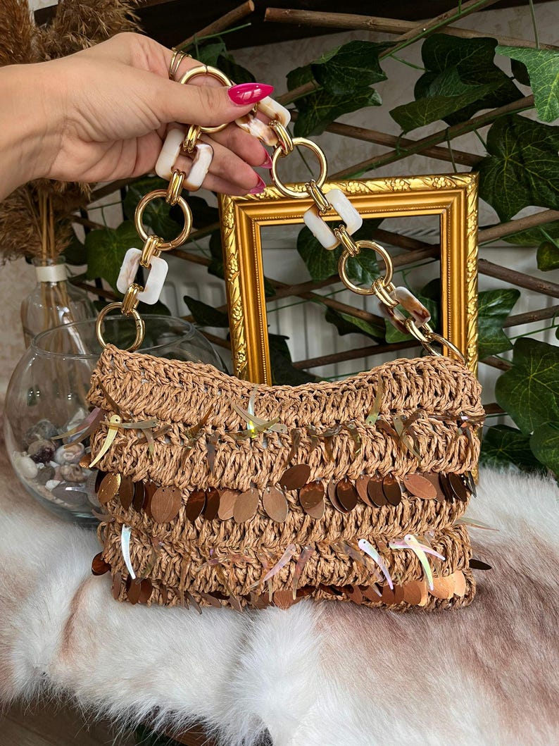 K&ouml;nnte beinhalten: Eine gewebte Raffia-Handtasche mit einem dekorativen Fransenbesatz aus Pailletten. Die Tasche hat einen goldfarbenen Kettenhenkel mit wei&szlig;en und braun marmorierten Akzenten. Die Tasche wird von einer Hand gehalten.