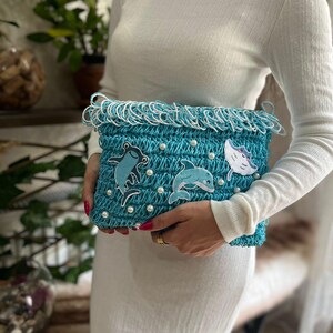 May include: A turquoise crocheted clutch with white fringe and pearl accents. The clutch features appliqued sea creature designs, including a frog, a dolphin, and a fish. The bag is held by a person wearing a white long-sleeved shirt and dress.