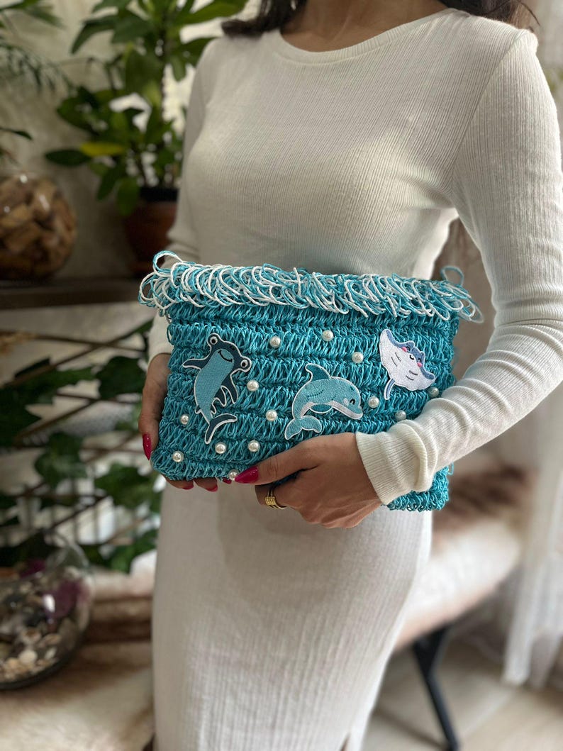 May include: A turquoise crocheted clutch with white fringe and pearl accents. The clutch features appliqued sea creature designs, including a frog, a dolphin, and a fish. The bag is held by a person wearing a white long-sleeved shirt and dress.