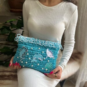 May include: A turquoise crochet clutch with white fringe and pearl accents. The clutch features embroidered sea creatures, including a dolphin, a stingray, and a seashell. The bag is held by a person wearing a white dress.