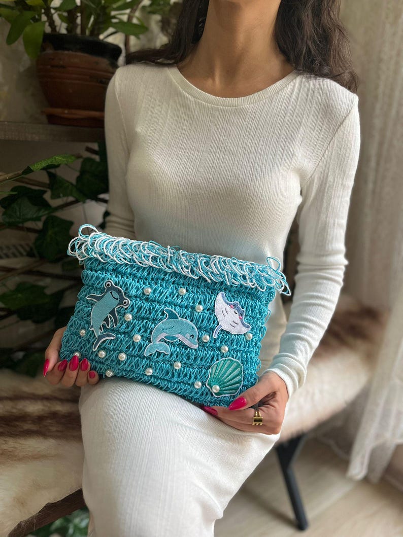 May include: A turquoise crochet clutch with white fringe and pearl accents. The clutch features embroidered sea creatures, including a dolphin, a stingray, and a seashell. The bag is held by a person wearing a white dress.