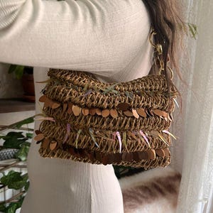 K&ouml;nnte beinhalten: Eine gewebte braune Handtasche mit goldenen Kettenriemen und dekorativen Pailletten. Die Tasche hat eine rechteckige Form und besteht aus einem Naturfasermaterial. Die Pailletten sind in Braun- und Regenbogenfarben gehalten.