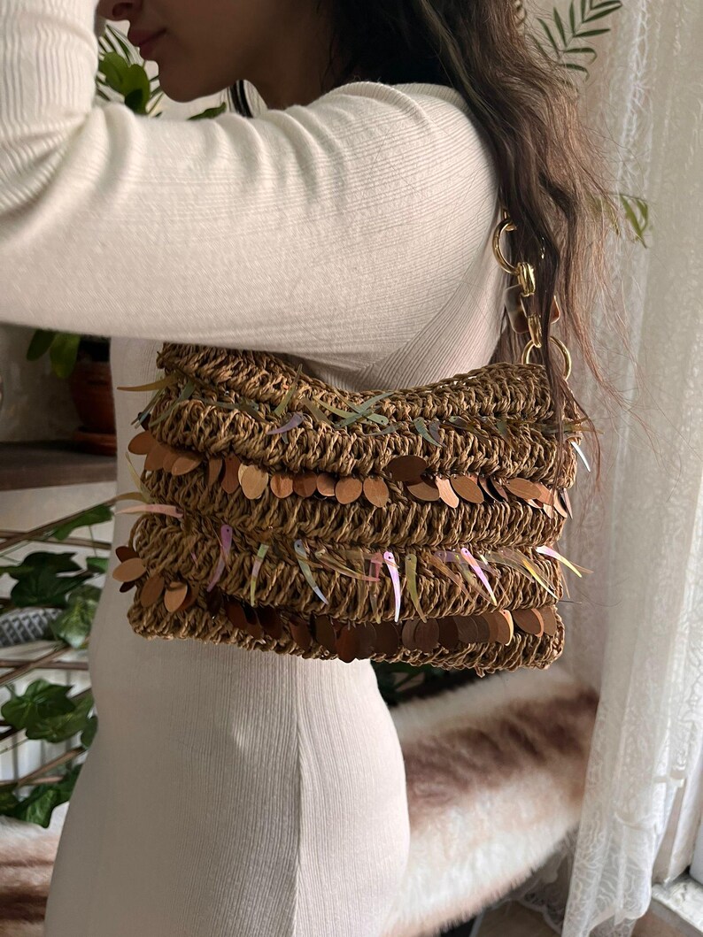 K&ouml;nnte beinhalten: Eine gewebte braune Handtasche mit goldenen Kettenriemen und dekorativen Pailletten. Die Tasche hat eine rechteckige Form und besteht aus einem Naturfasermaterial. Die Pailletten sind in Braun- und Regenbogenfarben gehalten.