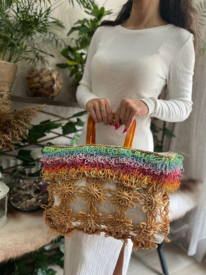 Puede incluir: Un bolso tote hecho a mano con una parte superior de color arco&iacute;ris y un dise&ntilde;o floral. El bolso tiene un interior blanco y asas marrones. El bolso est&aacute; hecho de materiales tejidos.