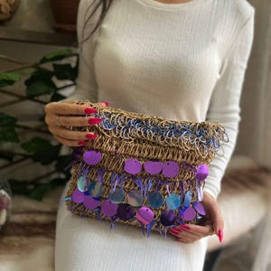 May include: A woven clutch with a brown base, blue trim, and purple sequin accents. The clutch is rectangular and has a fringed edge. The sequins are circular and iridescent, reflecting shades of purple and blue.