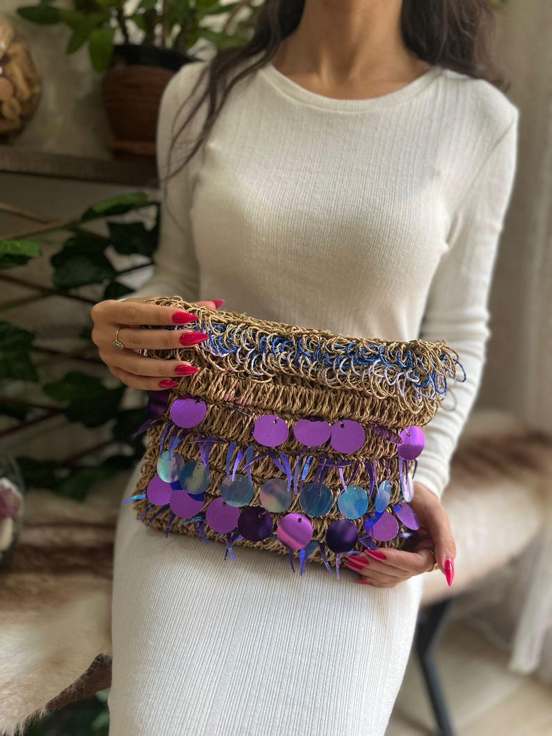 May include: A woven clutch with a brown base, blue trim, and purple sequin accents. The clutch is rectangular and has a fringed edge. The sequins are circular and iridescent, reflecting shades of purple and blue.