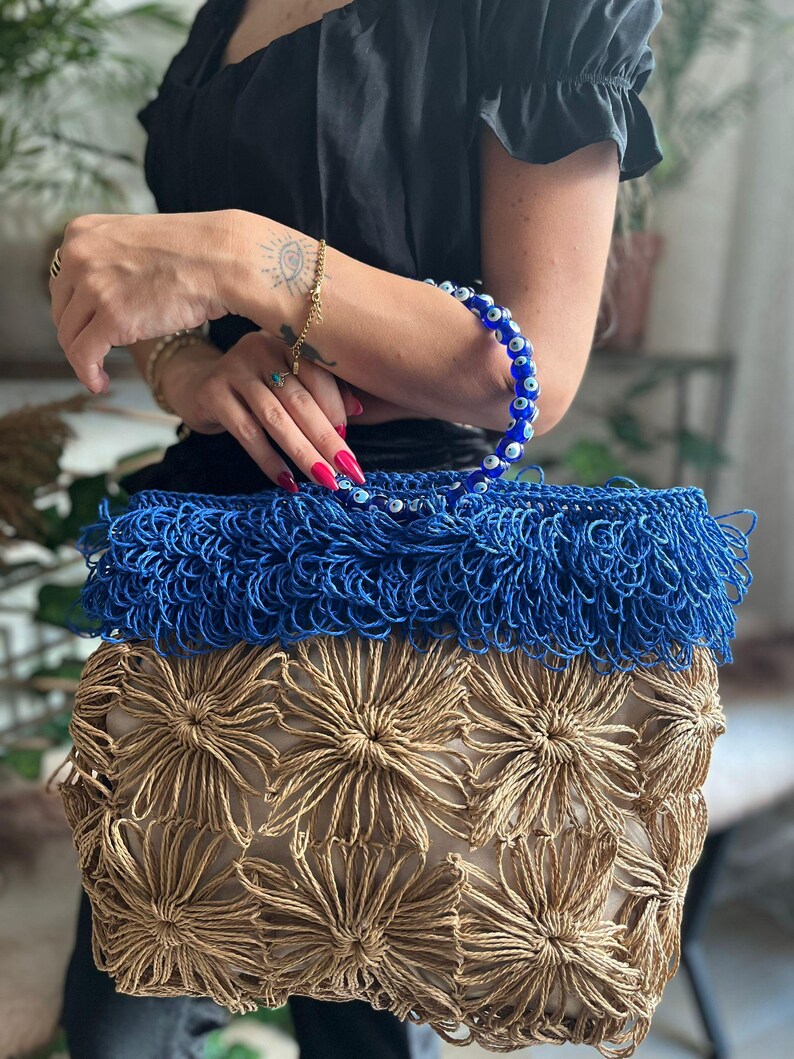 May include: A handmade tote bag with a blue fringe top and a beige floral design. The bag has a blue beaded handle. The bag is made of woven material.