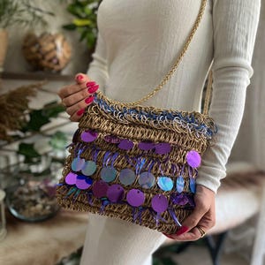 May include: A woven shoulder bag with a gold chain strap. The bag features a brown base with blue and gold trim, and is adorned with large, iridescent purple sequins. The bag is held by a person wearing a white long-sleeved shirt.