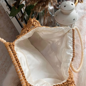 May include: Open view of a woven straw handbag with a cream-colored fabric lining. The bag has a beaded handle and a decorative fringe along the top edge. The bag is a light brown color.