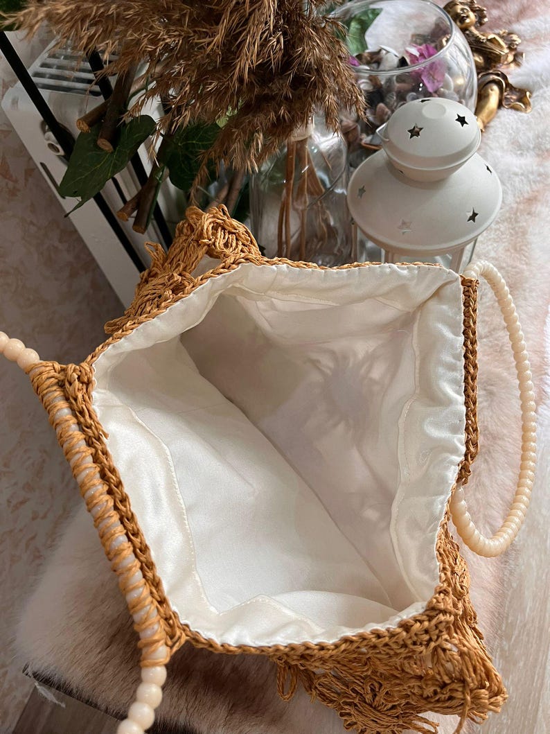 May include: Open view of a woven straw handbag with a cream-colored fabric lining. The bag has a beaded handle and a decorative fringe along the top edge. The bag is a light brown color.