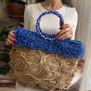 May include: A handmade tote bag with a blue handle adorned with blue and white beads. The bag features a woven blue top section and a lower section with a tan floral design. The bag is held by a person.