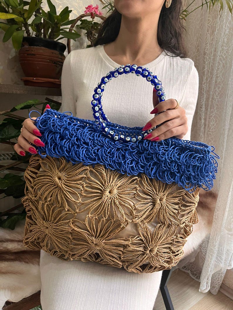 May include: A handmade tote bag with a blue handle adorned with blue and white beads. The bag features a woven blue top section and a lower section with a tan floral design. The bag is held by a person.