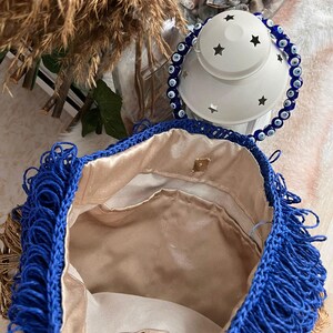 May include: A handmade blue crochet bag with fringe detail. The bag has a gold-colored clasp and a beige satin lining. The bag is open, revealing the interior. A white lantern with star cutouts and a blue beaded bracelet are in the background.