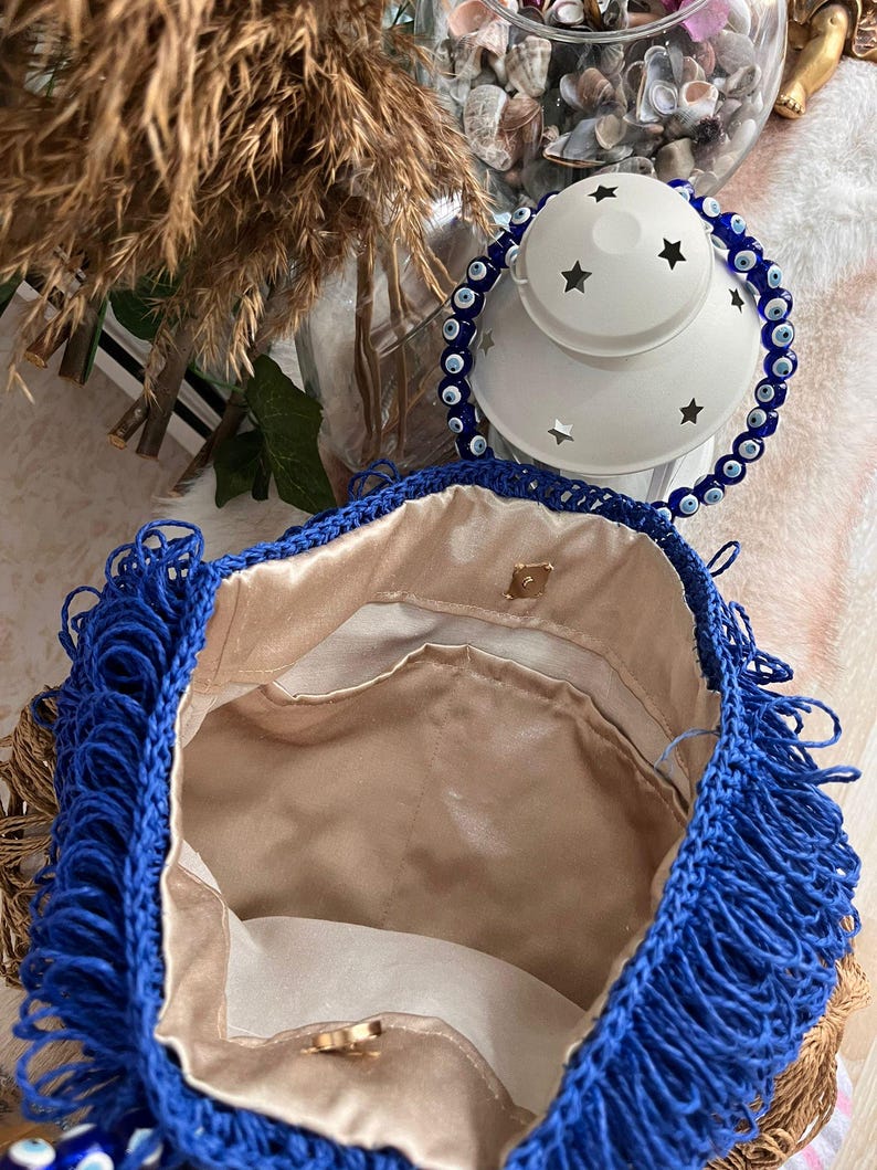 May include: A handmade blue crochet bag with fringe detail. The bag has a gold-colored clasp and a beige satin lining. The bag is open, revealing the interior. A white lantern with star cutouts and a blue beaded bracelet are in the background.