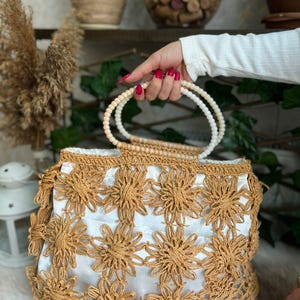 May include: A woven handbag with a white interior and a floral design in a light brown color. The bag has a beaded handle and a fringe detail along the bottom edge. The bag is held by a person.