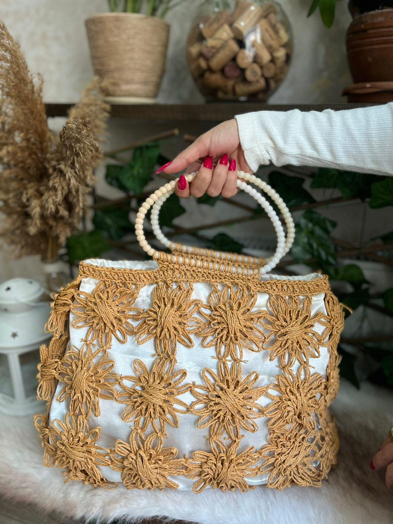 May include: A woven handbag with a white interior and a floral design in a light brown color. The bag has a beaded handle and a fringe detail along the bottom edge. The bag is held by a person.