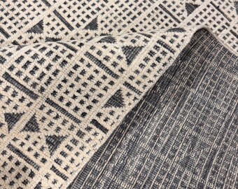 May include: A rolled-up area rug with a geometric pattern. The rug features a repeating design of dark grey triangles and squares on a cream-coloured background. The underside of the rug is a solid dark grey colour.