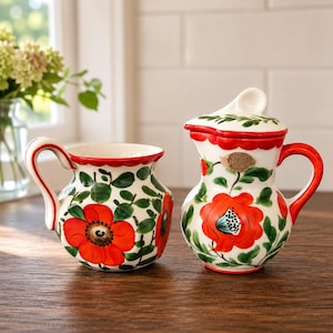 May include: A ceramic creamer and a lidded sugar bowl featuring a hand-painted pattern of vibrant red flowers and green leaves on a cream-coloured background. The sugar bowl has a small metal plaque with the text "ZEPHILA" on the front.
