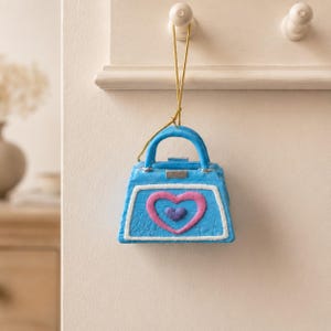 Op de afbeelding: Een blauw handtasornament met een textuurrijk oppervlak, voorzien van een witte rand, een roze hartomlijning en een klein paars hartje in het midden. De handtas heeft een handvat en hangt aan een dun gouden koordje voor ophangdoeleinden.