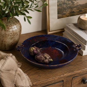 May include: A deep blue ceramic serving dish with a red interior, featuring two artichokes. The dish has decorative handles and is placed on a wooden surface, alongside a vase and a framed picture.