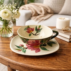 May include: A cream-colored teacup and saucer set with a floral design in pink and green. The teacup has a dark green handle and sits on a matching saucer. The set is on a wooden table, with a vase of flowers and a candle in the background.