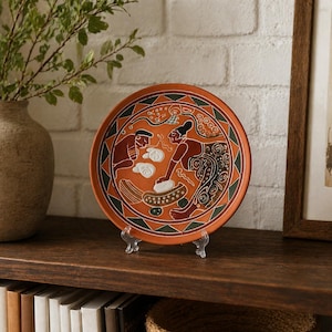 May include: A terracotta-colored decorative plate with a detailed illustration of two figures. The plate features a geometric border with green and black triangles. The figures are depicted in brown and black, with white cloud-like shapes. The plate is displayed on a clear stand.