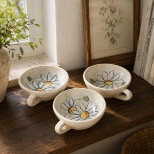 May include: Three off-white ceramic bowls with a single handle, each featuring a hand-painted blue and yellow daisy flower design on the interior. The bowls have a rounded shape and a smooth, glossy finish, showcasing a consistent floral pattern.