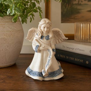 Puede incluir: Una figura de ángel de color crema con cabello largo y alas emplumadas. El ángel viste una túnica con adornos azules en el cuello, los puños y el dobladillo. Un pergamino cruza la parte delantera de la túnica con el texto Special teacher.