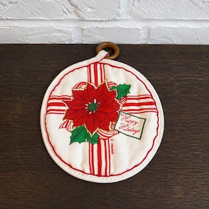 May include: A round, off-white quilted potholder featuring a red poinsettia flower with green leaves, red and white striped ribbon patterns, and a small tag with the text Happy Holidays. The item includes a brown wooden ring for hanging.