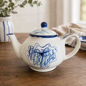 May include: A white ceramic teapot featuring a hand-painted blue and green abstract scribble design on the body and lid. The teapot has a curved handle, a tapered spout, and a matching lid with a small round knob, all accented with blue trim.