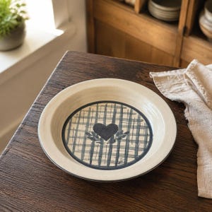 May include: A round ceramic plate with a cream-colored rim and a central design featuring a dark blue painted heart surrounded by leafy sprigs, all set against a dark blue and cream-colored plaid pattern on the interior base of the dish.