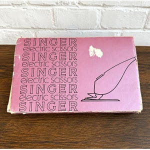May include: A rectangular cardboard box for Singer electric scissors with a mauve background. The text SINGER electric scissors is printed repeatedly in black on the left side. A black line drawing of the electric scissors is on the right.