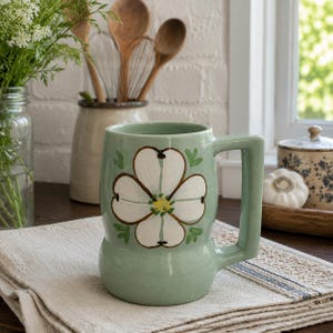 May include: A light green ceramic mug featuring a hand-painted white dogwood flower with a yellow center on the front. The mug has a wide, flat-sided handle and a rounded base, sitting on a folded beige linen cloth with a blue striped border.