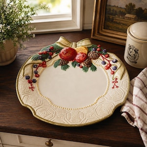 May include: A round ceramic serving plate with a cream-colored center and a wide, embossed geometric border. The top edge features a raised, hand-painted holiday motif of pine branches, red berries, pinecones, and a central cream-colored bow.