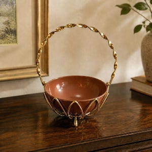 May include: A decorative basket featuring a smooth, terracotta-colored bowl held within a gold-toned metal frame. The frame includes a tall, twisted metal handle and a base constructed from curved, petal-like metal supports that cradle the bowl.