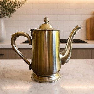 May include: A metallic, brass-toned teapot with a cylindrical body, a curved handle, a long tapered spout, and a small finial on the lid. The teapot features a smooth, polished finish and a wide, stable base, presenting a classic, functional design.
