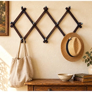 May include: An expandable dark brown wooden wall rack with a diamond-shaped lattice design and multiple pegs. A beige fabric tote bag hangs from one peg, and a tan straw hat with a black ribbon band hangs from another peg on the wall-mounted rack.