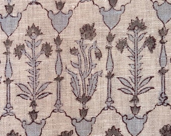 Op de afbeelding: Close-up van een beige stof met een herhalend patroon van gestileerde bloemen en geometrische vormen in gedempt blauw en bruin. Het ontwerp bevat verticale bloemelementen en boogmotieven, waardoor een gestructureerd, decoratief oppervlak ontstaat.