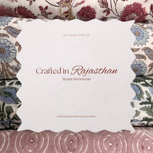 Op de afbeelding: Een witte kaart met een geschulpte rand toont de tekst "Crafted in Rajasthan, Styled Worldwide". De kaart bevindt zich voor textiel met bloemmotieven in roze, blauwe en witte tinten. De tekst "Hand block printed natural textile" is ook zichtbaar.