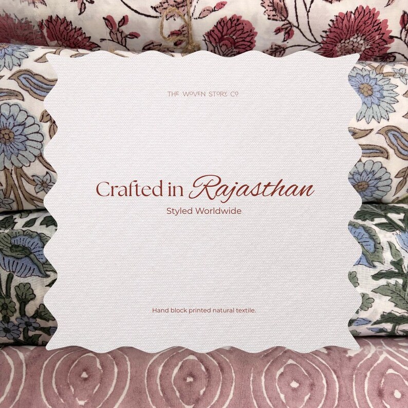 Op de afbeelding: Een witte kaart met een geschulpte rand toont de tekst "Crafted in Rajasthan, Styled Worldwide". De kaart bevindt zich voor textiel met bloemmotieven in roze, blauwe en witte tinten. De tekst "Hand block printed natural textile" is ook zichtbaar.