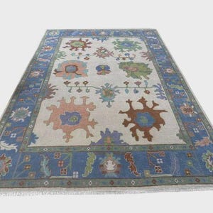 May include: Rectangular rug with a light beige central area and a blue border. The rug displays a repeating pattern of stylized floral and geometric designs in blue, green, orange, and purple. The rug has a fringe along the edges.