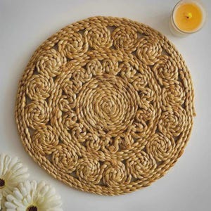 May include: A round, woven placemat made of natural fibers. The placemat features a spiral design in the center, surrounded by a repeating pattern of looped spirals. The color is a warm, light brown, and the texture appears slightly rough.